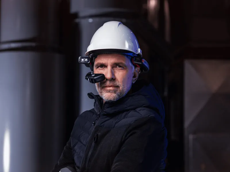 A technician in a white hard hat wearing a helmet-mounted heads-up display, in a dark plant room
