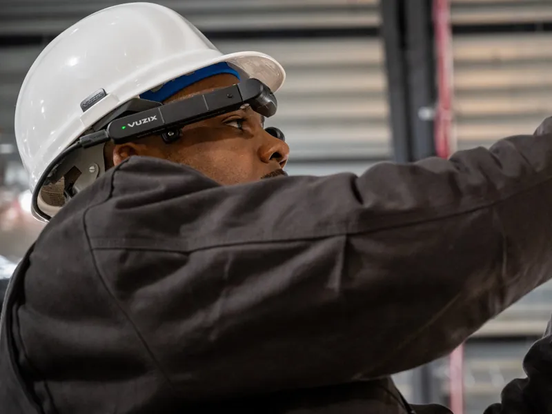 A technician in a white hard hat wearing hat-mounted smart glasses, reaching up to work overhead